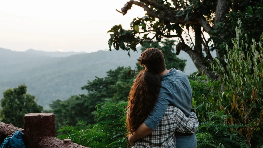Couples travel psychology: a couple embracing at a forest viewpoint, watching the sunset over misty green hills
