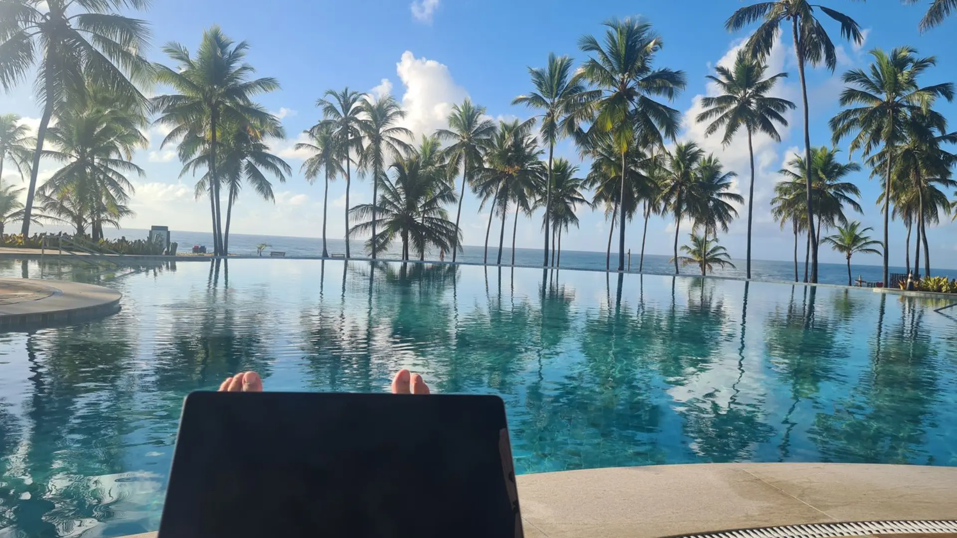 Laptop and bare feet by a resort infinity pool with palm trees and ocean view