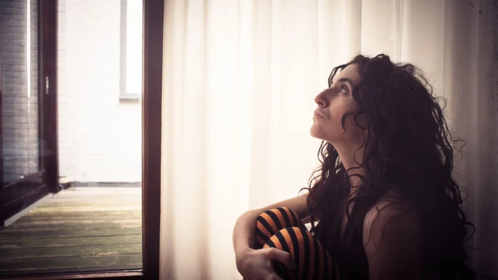 Woman sitting by a window hugging her knees and looking upward, lost in thought