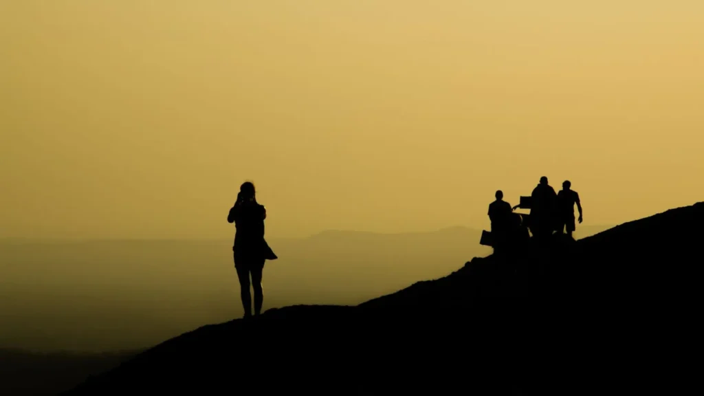 Silhouettes of travellers on a hilltop at sunset, one person photographing the view while a group walks along the ridge