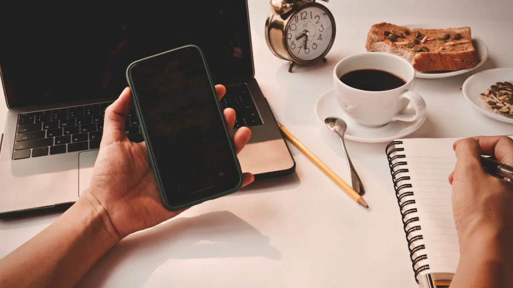 Hand writing in a spiral notebook beside coffee, toast and an alarm clock on a morning desk