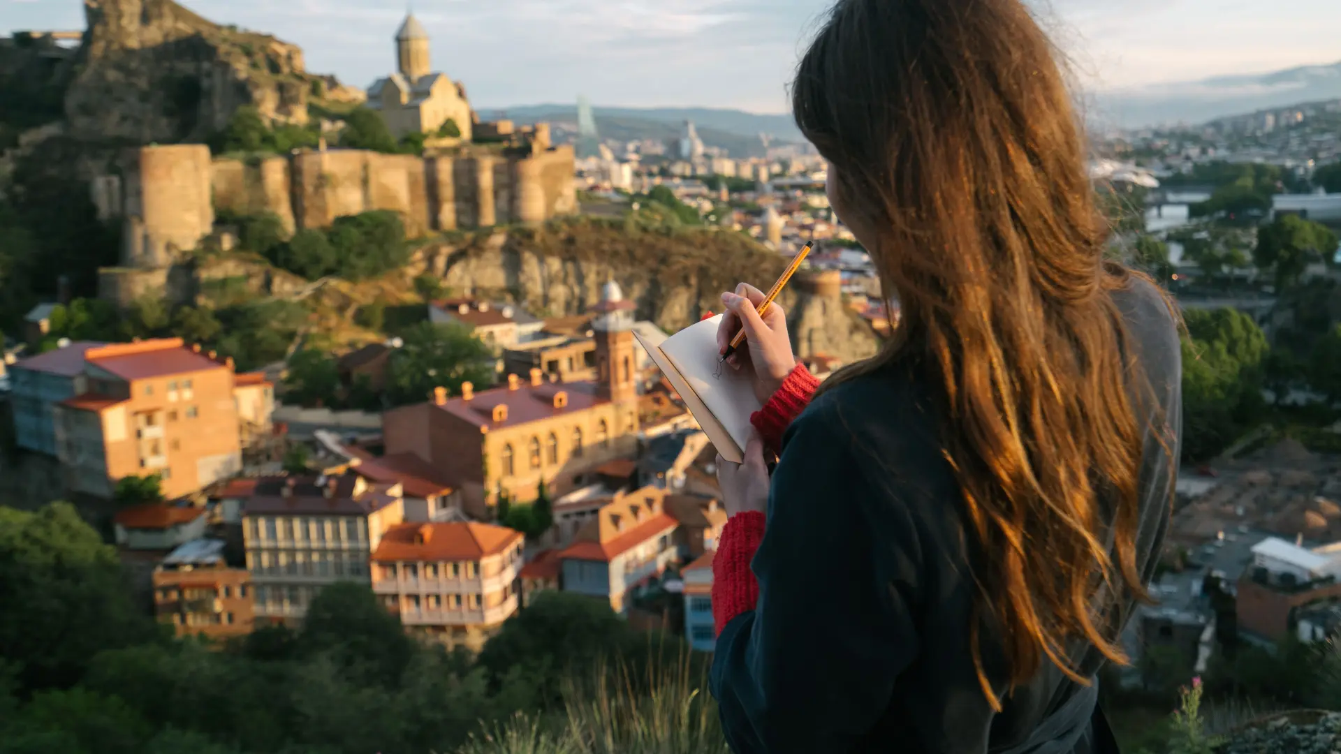 Woman writing in a travel journal on a hilltop overlooking an old city at sunset