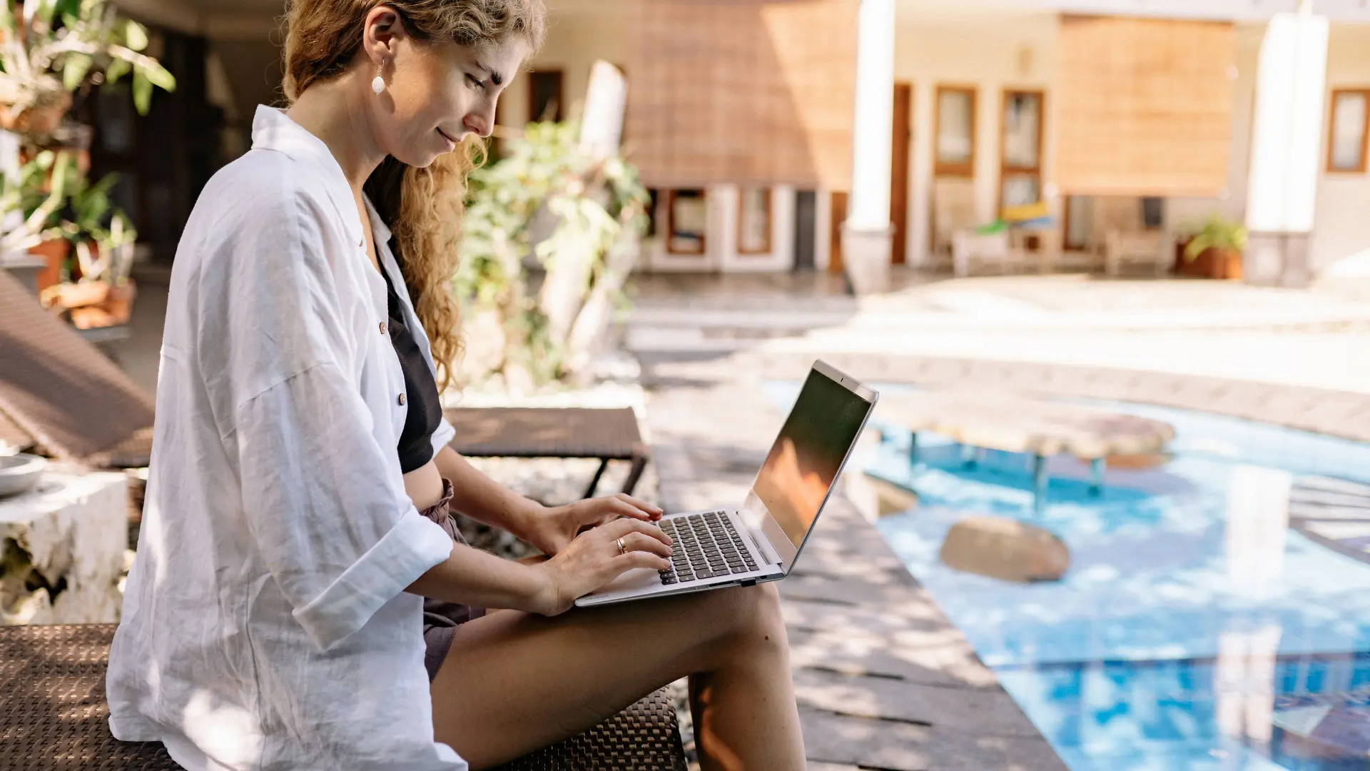 Relaxed woman typing on a laptop beside a resort pool, planning a trip calmly
